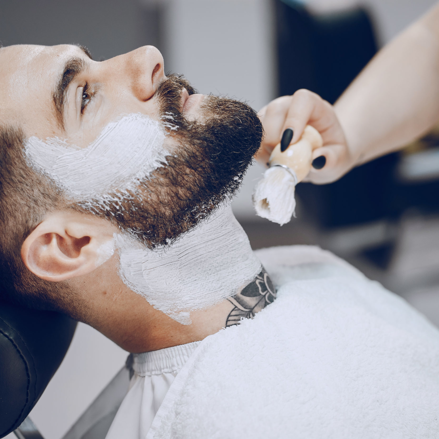 handsome young bearded guy sitting in an armchair in a beauty salon and the girl around him smears his beard with cream